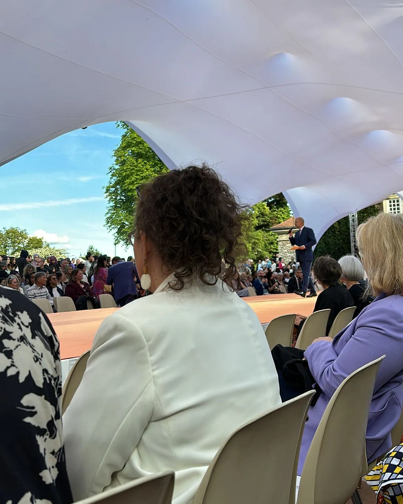 Public assis de dos sous une grande tente blanche pendant un défilé, un intervenant au micro sur le podium